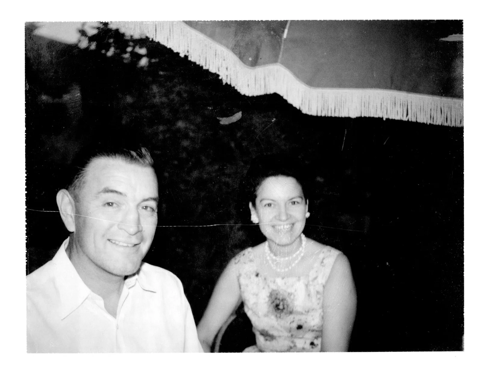Black and white photo of a smiling man and woman seated under a fringed umbrella.