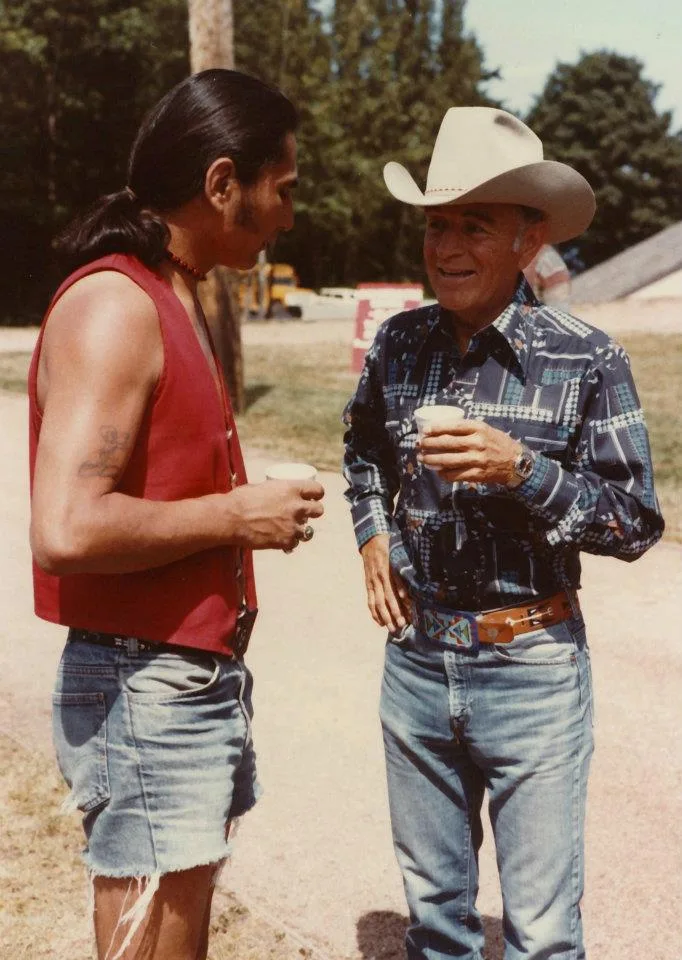 A young man with long hair in a red vest talking with an elder in a cowboy hat, both holding cups.