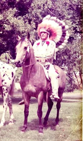 A rider in a full feathered headdress and white buckskin seated on a horse on a lawn.