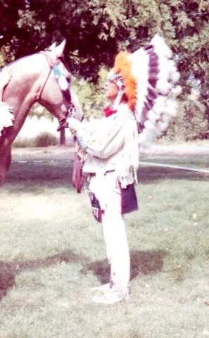 A person in a feathered headdress and white fringed buckskin standing beside a horse on a lawn.
