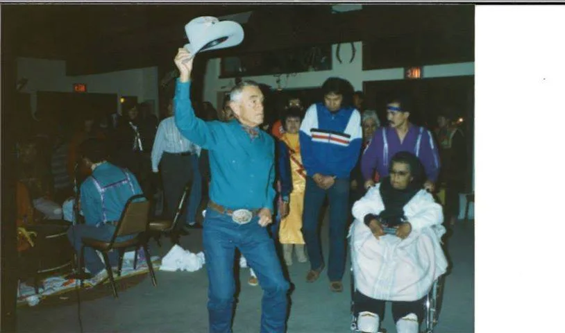 A man raising his cowboy hat while people walk behind him at an indoor gathering, one person in a wheelchair.