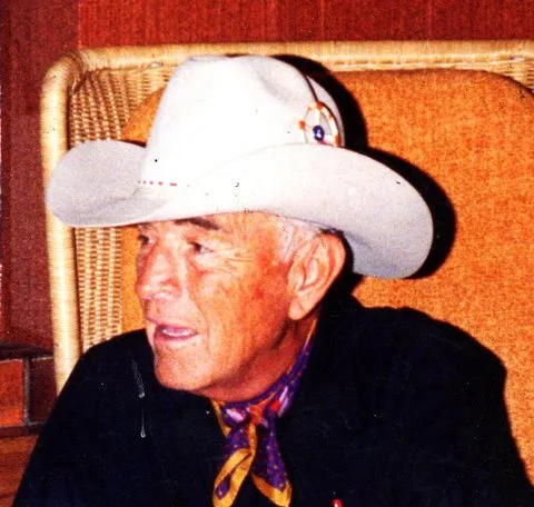 Close-up of an elder in a white cowboy hat and patterned scarf, seated in a wicker chair.