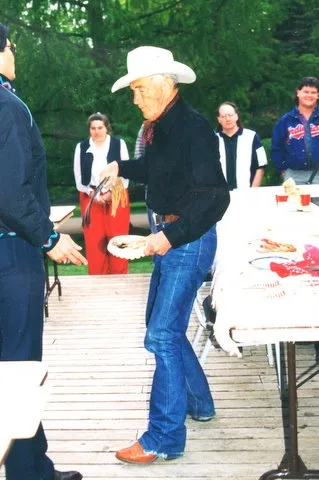 An elder in a cowboy hat holding a plate at an outdoor buffet table on a wooden deck.