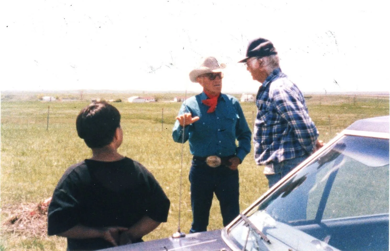 An elder in a cowboy hat and red bandana talking with a man in a plaid shirt and a boy on open prairie beside a car.