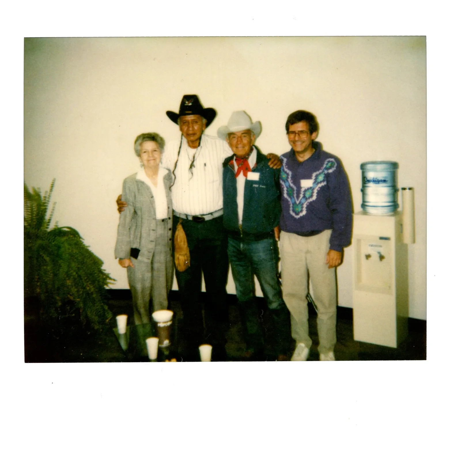 Polaroid of four people: a woman, a man in a black hat with braids, an elder in a white cowboy hat and red bandana, and a man in a purple sweater.