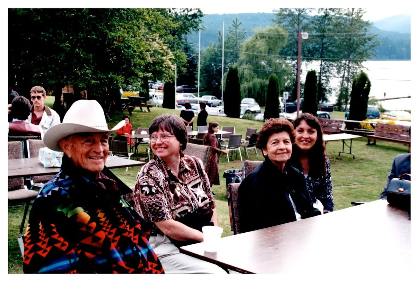 An elder in a white cowboy hat and patterned jacket seated at an outdoor table with three smiling women near a lake.