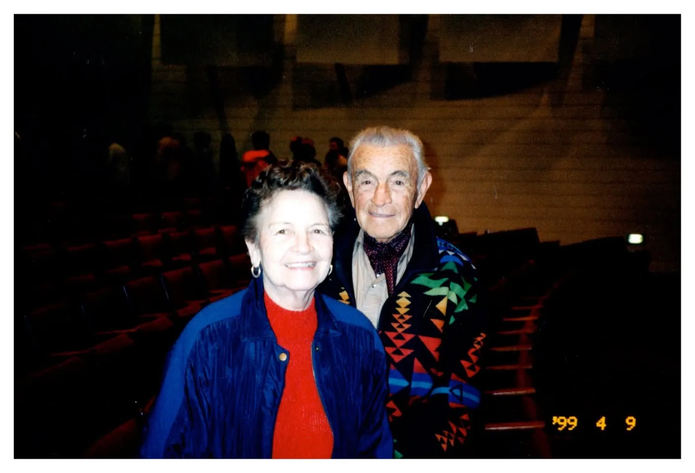 A smiling older couple standing in an auditorium, dated 1999.