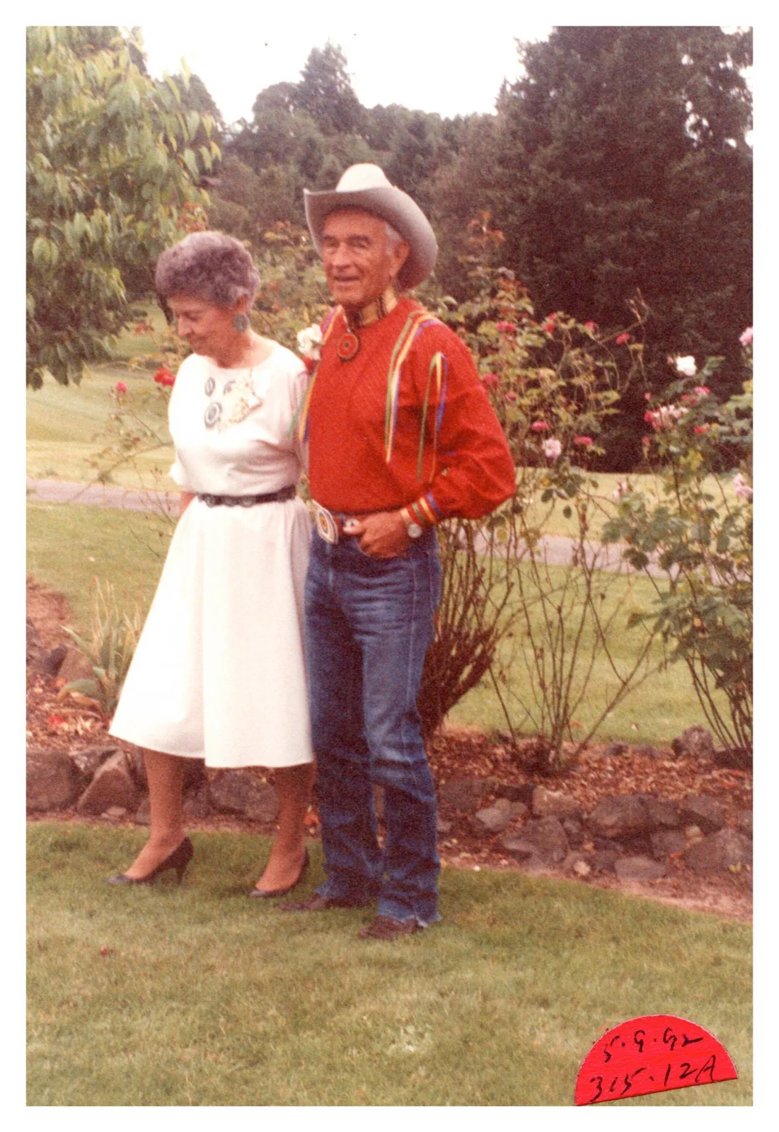 An older couple in a garden, the woman in a white dress and the man in a red ribbon shirt and cowboy hat.