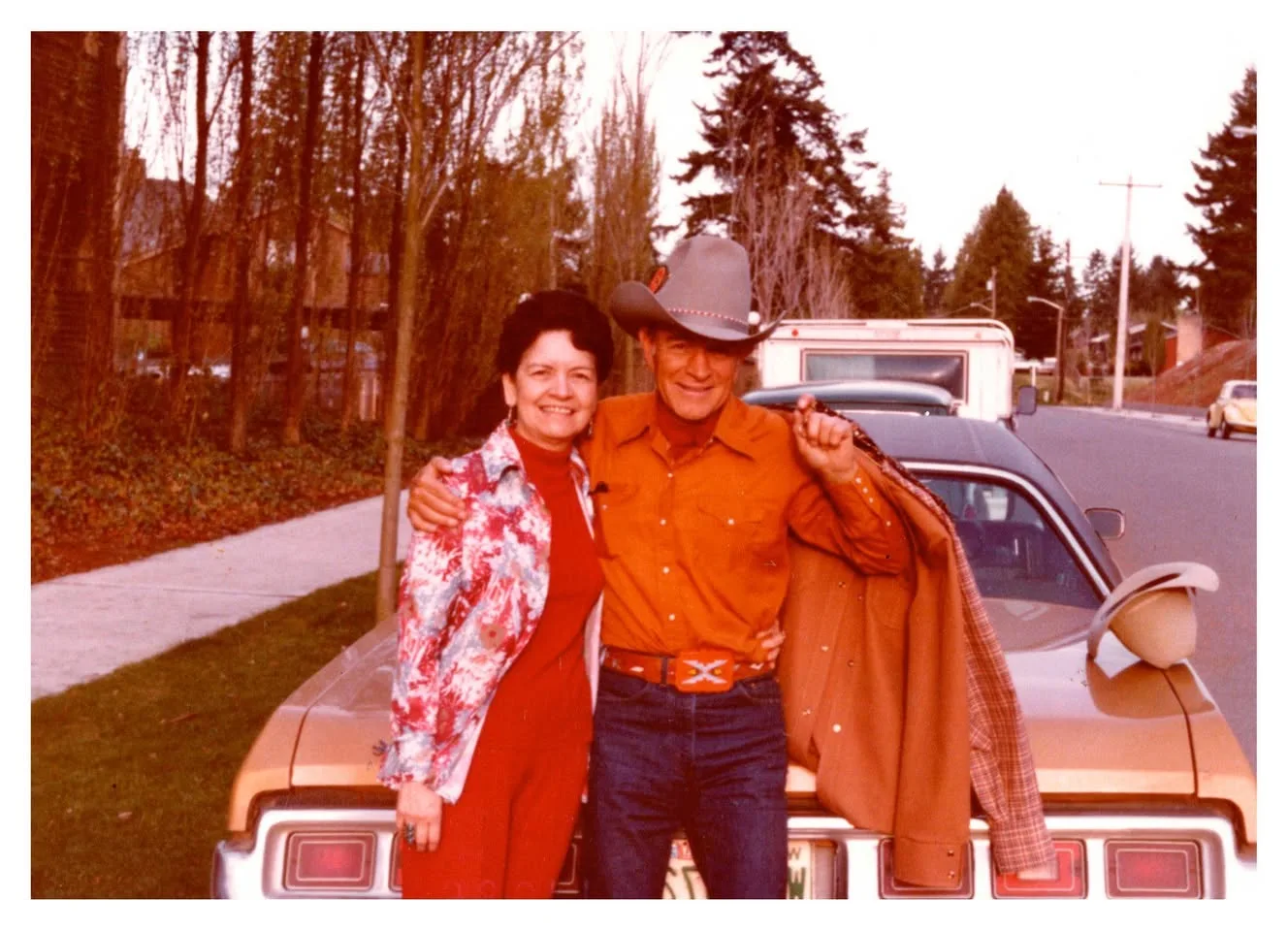 A smiling couple embracing beside a car, the man in an orange western shirt and cowboy hat.