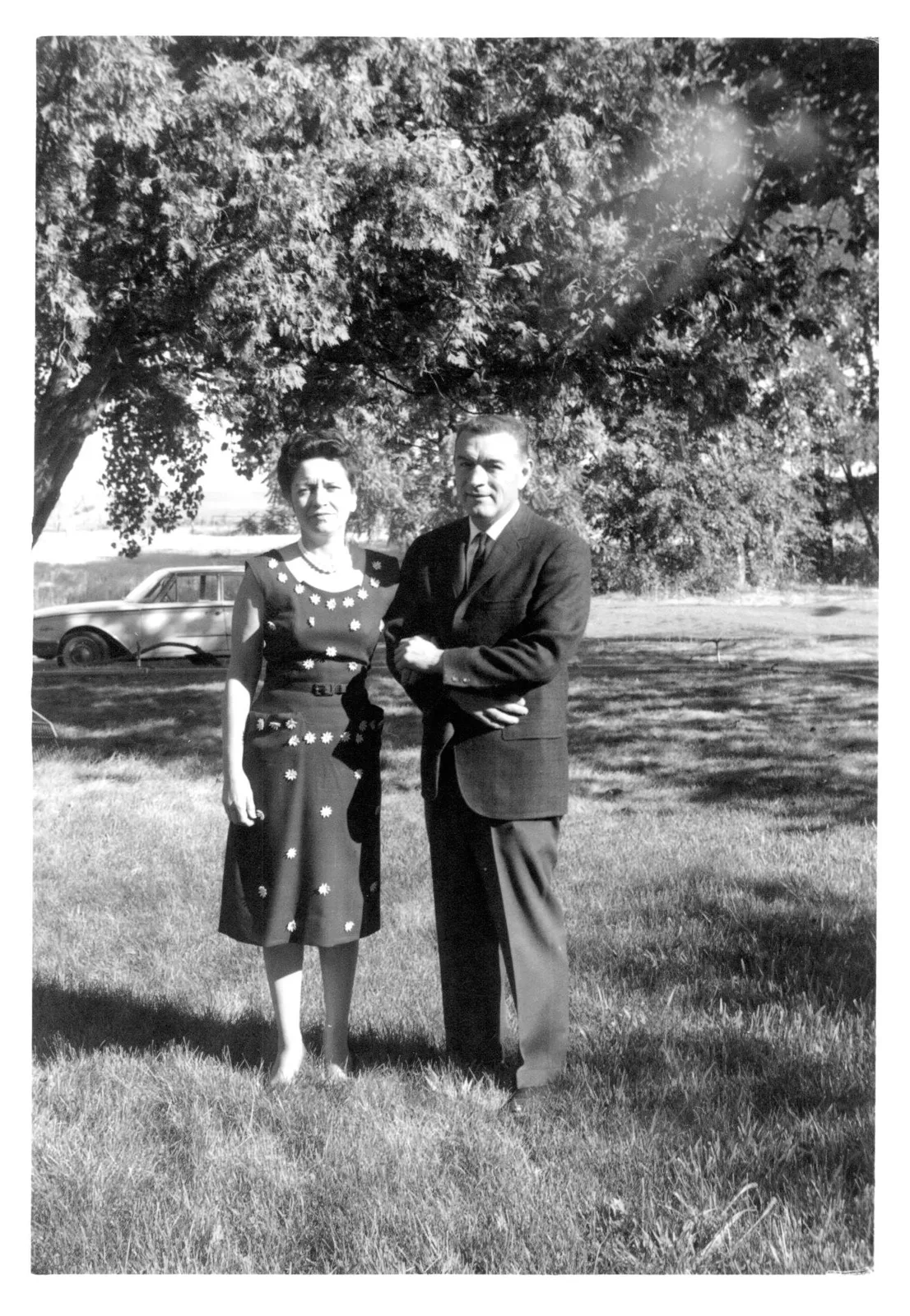 Black and white photo of a couple standing arm in arm on grass beneath a large tree.