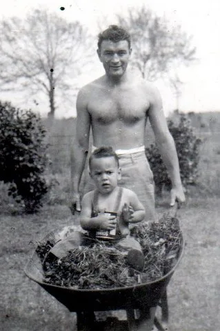 Vintage photo of a shirtless young man pushing a toddler in a wheelbarrow of leaves.