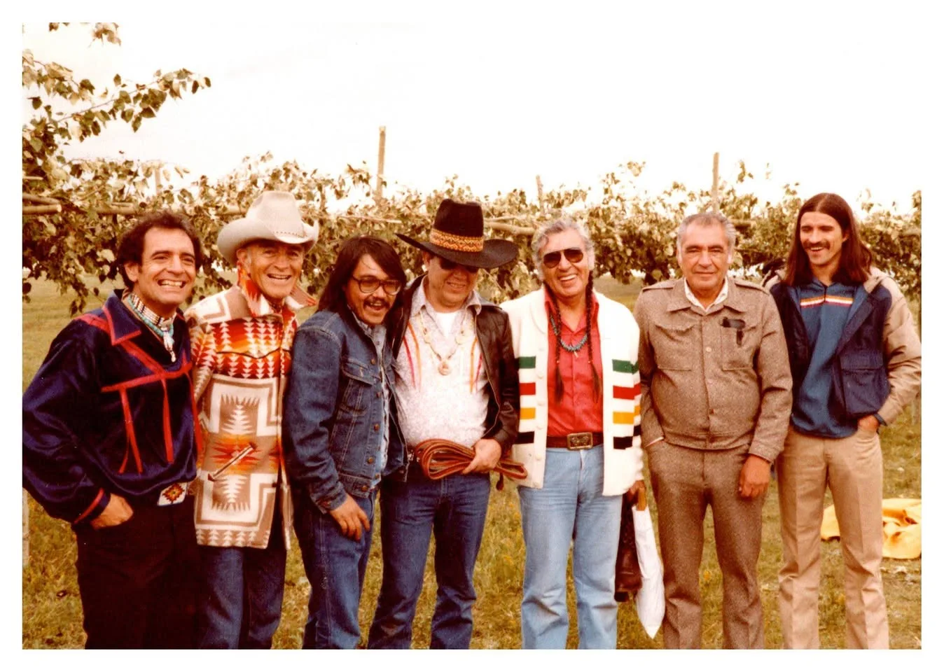 Seven smiling men standing outdoors in front of a row of young trees, one in a cowboy hat and patterned jacket.