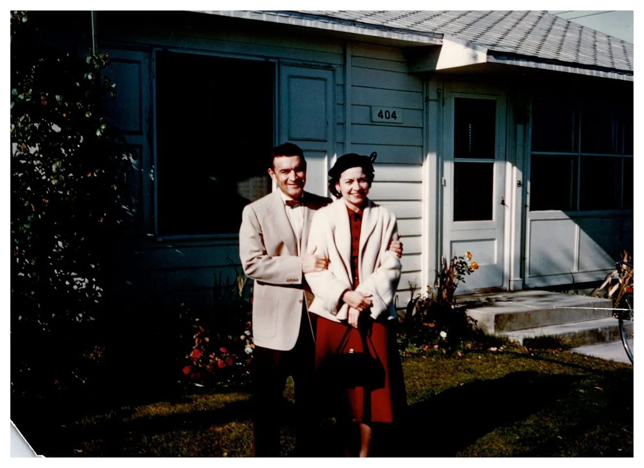 A smiling couple in front of a house numbered 404, the man in a light jacket and bow tie, the woman in a white coat and red dress.