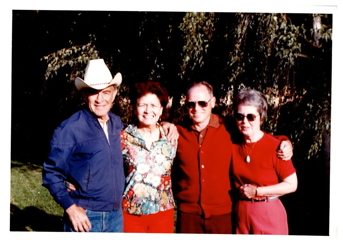Four older adults standing arm in arm before trees, one man wearing a white cowboy hat.