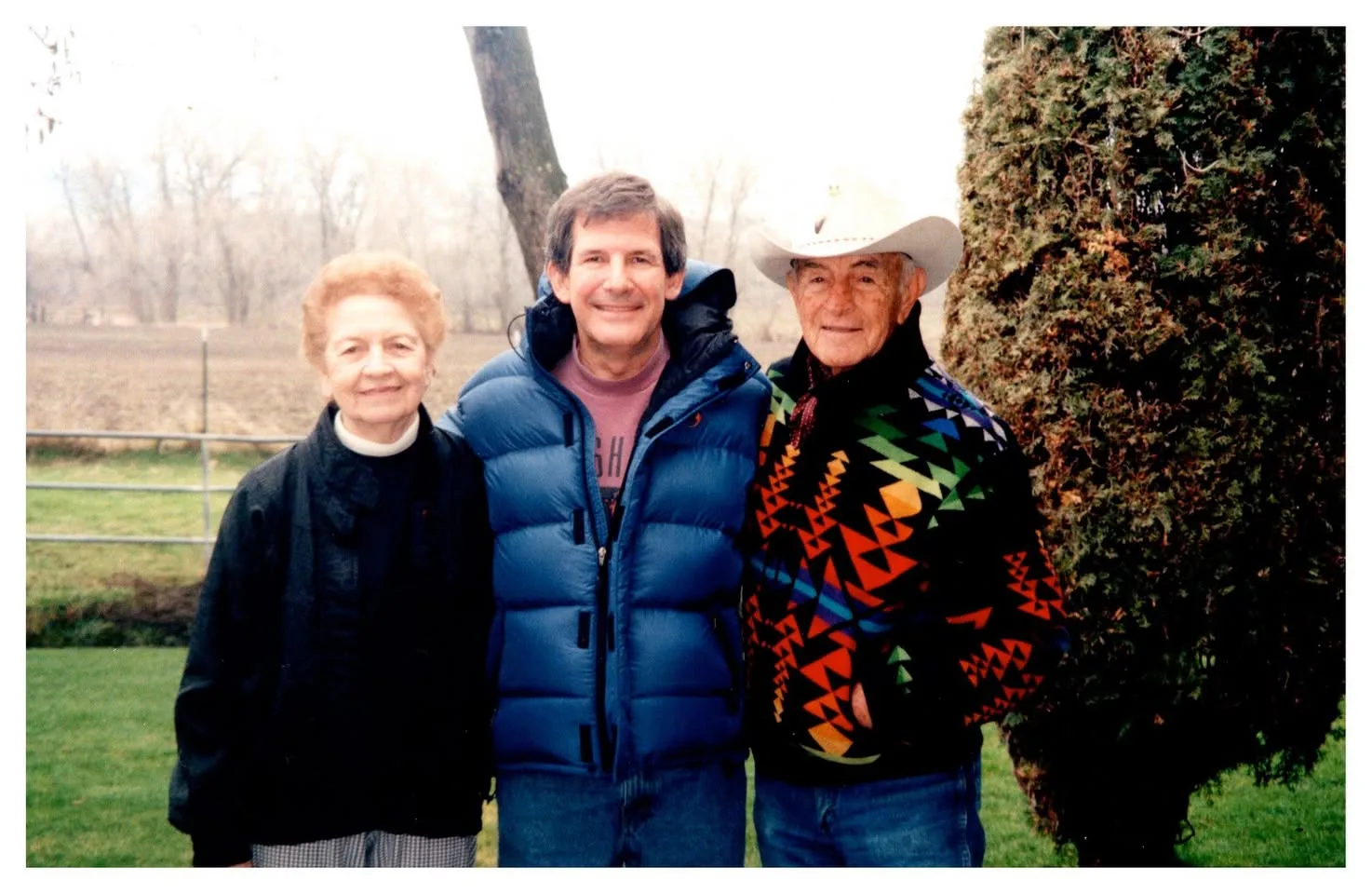 An older woman, a man in a blue puffer jacket and an elder in a white cowboy hat and patterned jacket standing outdoors.
