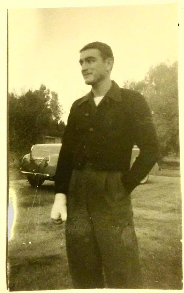 Old sepia photograph of Phil Lane Jr.'s father as a young man in 1940, standing on grass in a dark jacket with a light-colored right hand, a vintage car and trees behind him.