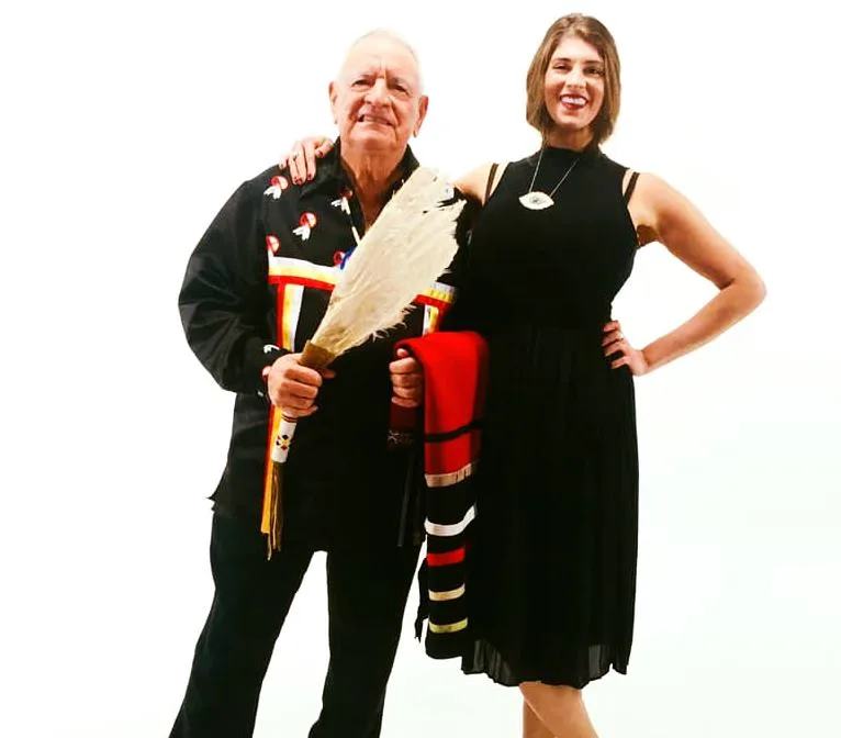 Phil Lane Jr., in a black ribbon shirt and holding a white feather fan, stands smiling with Toni Lane, who wears a black dress and holds a red striped blanket.