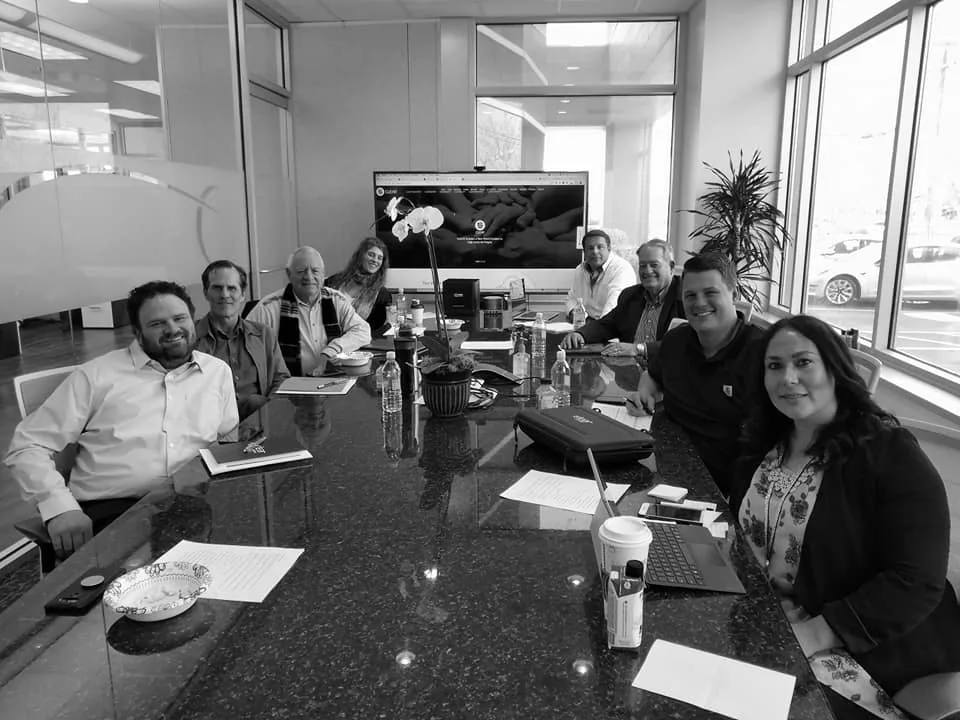 Black-and-white group of nine people seated around a long granite conference table with papers and water bottles, a large screen behind them.