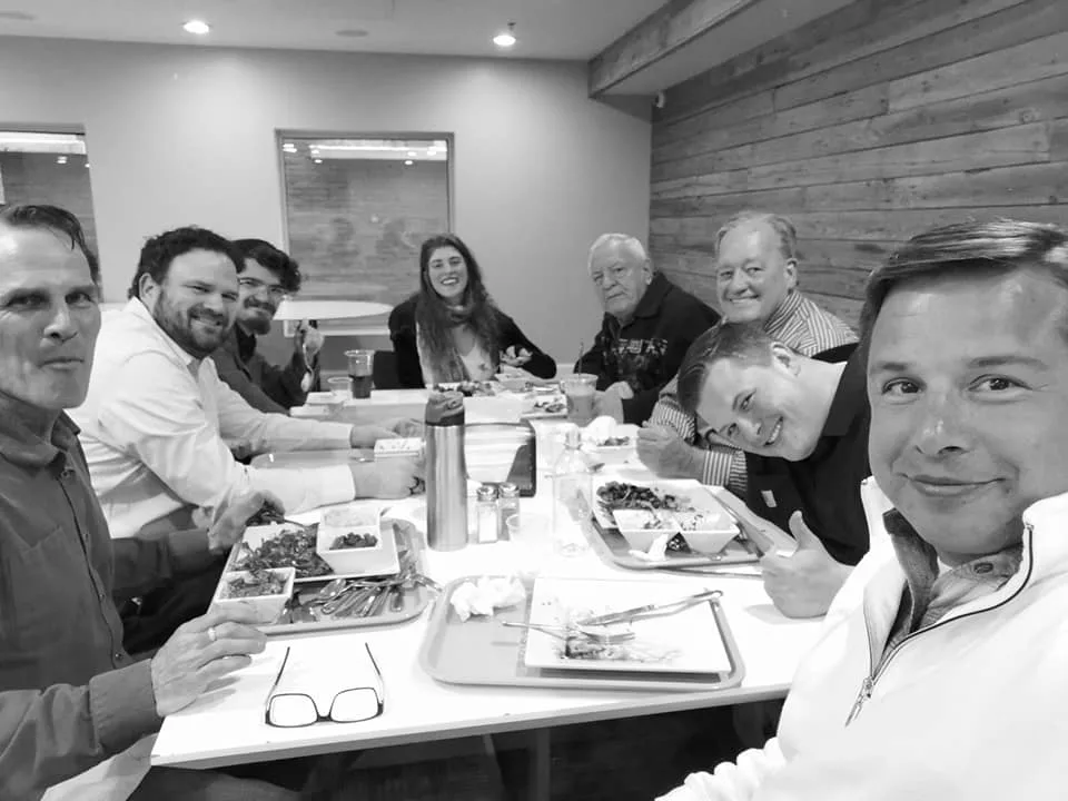 Black-and-white selfie of eight smiling people sharing a meal at a restaurant table with trays of food.