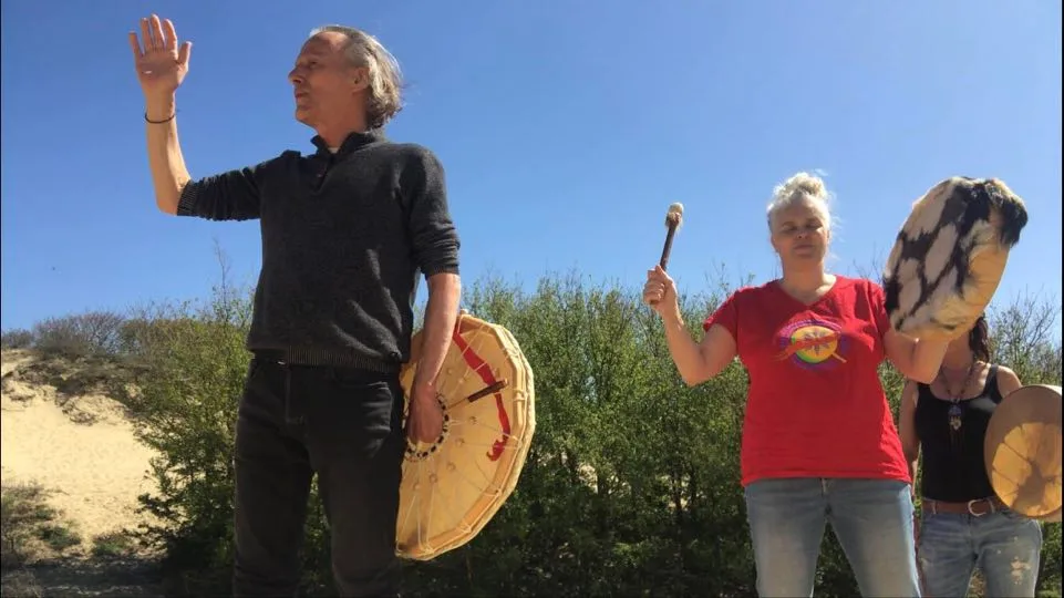 A man holding a hand drum raises his open palm, beside a woman in a red shirt lifting a drum and beater, with another drummer behind them, among green shrubs and sand dunes under a blue sky.