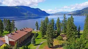 Aerial view of a resort lodge among tall evergreen trees on the shore of a wide blue lake ringed by forested mountains.