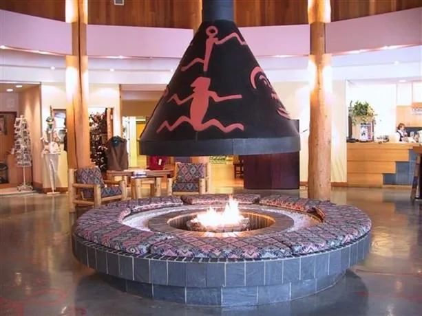 A hotel lobby with a round sunken fire pit and cushioned seating beneath a black conical hood painted with red pictograph-style figures.