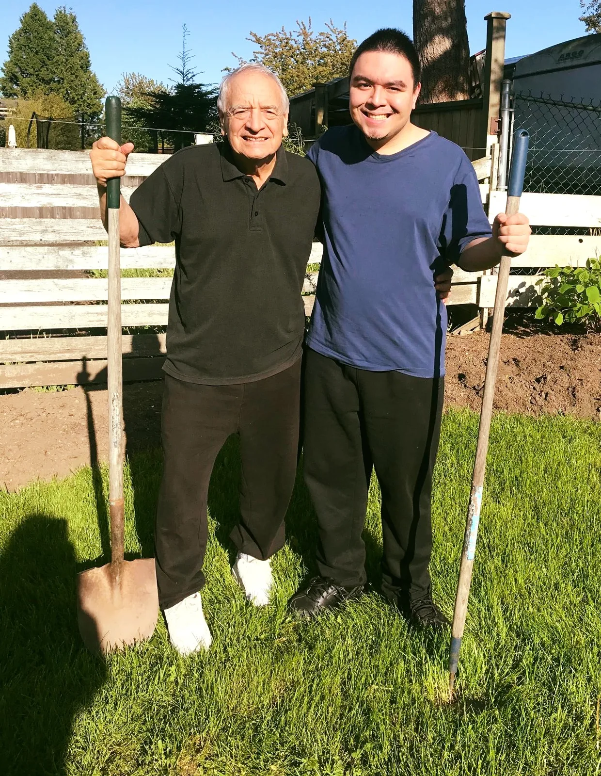 An elderly man in a black polo shirt holding a shovel stands smiling with his arm around a young man in a blue T-shirt holding a long-handled tool, on a sunny lawn by a garden and wooden fence; a small shadow falls at bottom left.