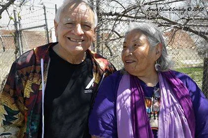 Phil Lane Jr., in a patterned jacket over a black shirt, smiles beside Tanksi Faith, a gray-haired woman in purple with a lavender scarf, by a chain-link fence and bare trees.