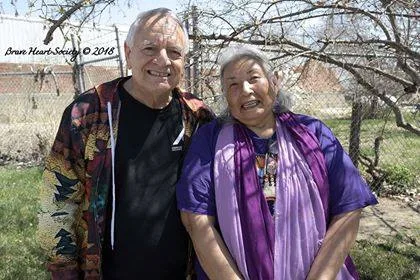 Phil Lane Jr. and Tanksi Faith stand side by side smiling outdoors on a sunny day, with a chain-link fence, bare trees and green grass behind them.