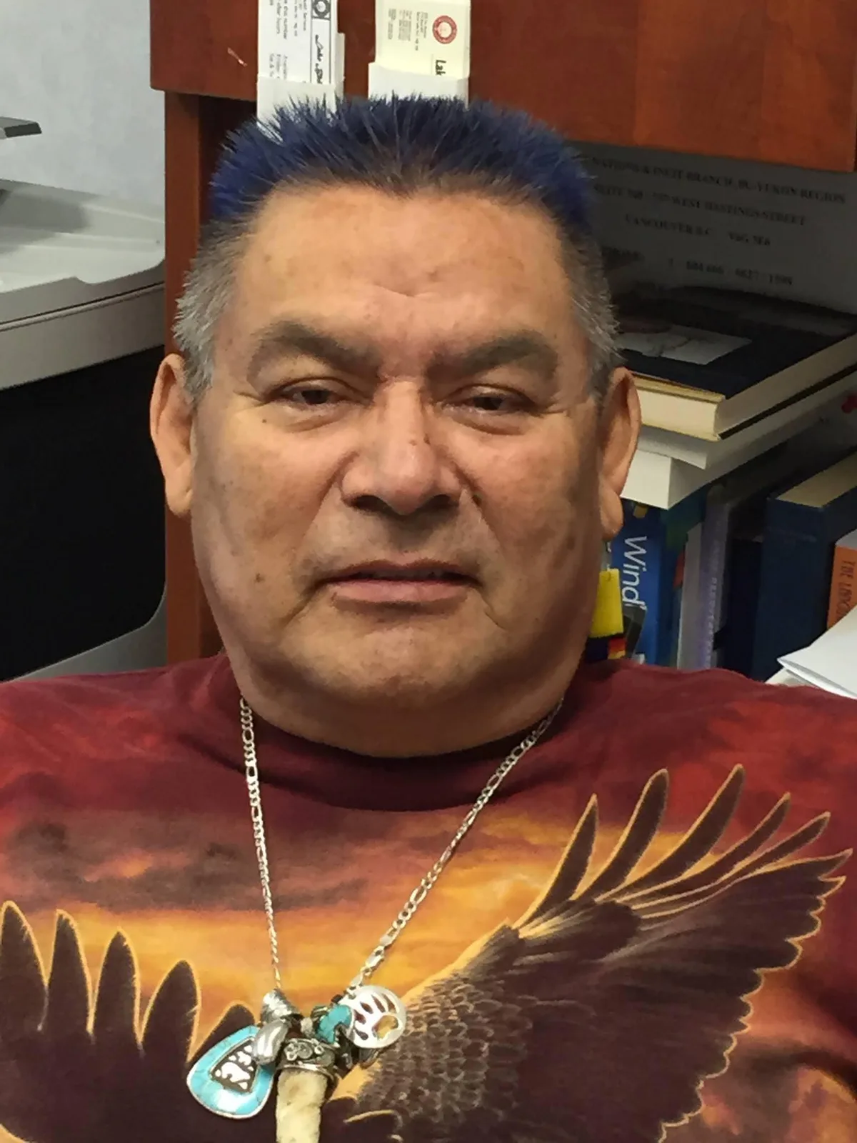 Howard Vincent, with spiky blue-tipped hair, wearing a red eagle T-shirt and a silver necklace with turquoise pendants, seated in an office.