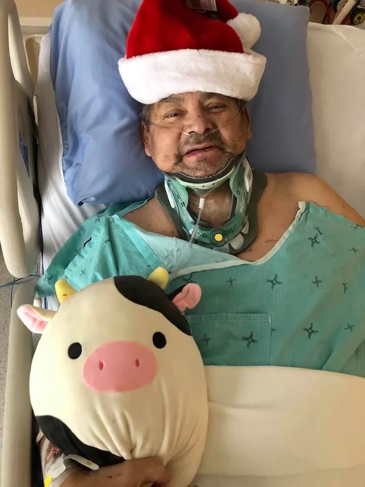 Howard Vincent smiling in a hospital bed, wearing a Santa hat, a neck brace and hospital gown, hugging a plush cow toy.