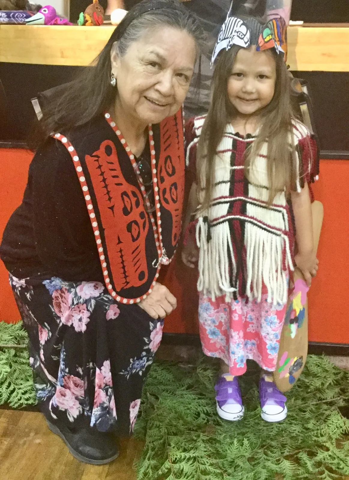 An Indigenous elder in a black and red button-trimmed vest with a floral skirt kneels beside a smiling young girl in a woven fringed top and paper headband, on cedar boughs.