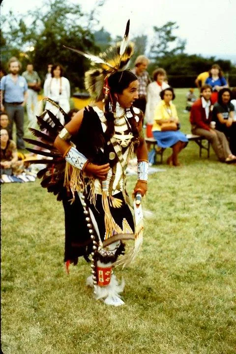Tahansi Kevin Isgor-Locke dances on a grassy field in black and white regalia with a feather roach and bustle, as a seated crowd watches behind him.