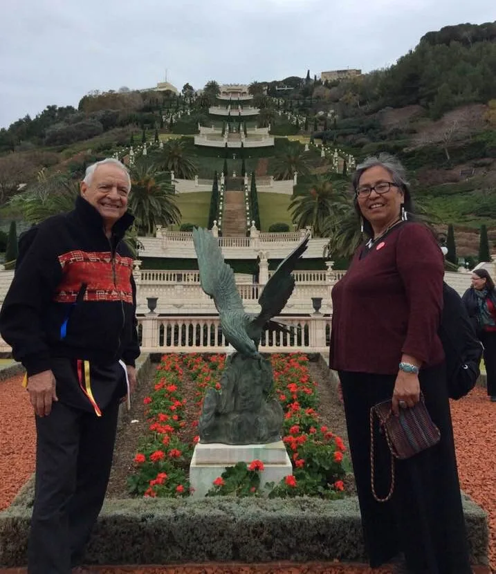 An elderly man in a black jacket and a woman in a maroon top stand beside a bronze eagle statue amid red flowers, with terraced gardens rising up a hillside behind.