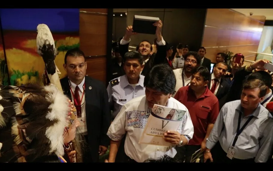 Evo Morales, in a white embroidered shirt, holds a booklet titled "V Cumbre Abya Yala" amid a crowd of officials and photographers, beside an elder in a feathered headdress.