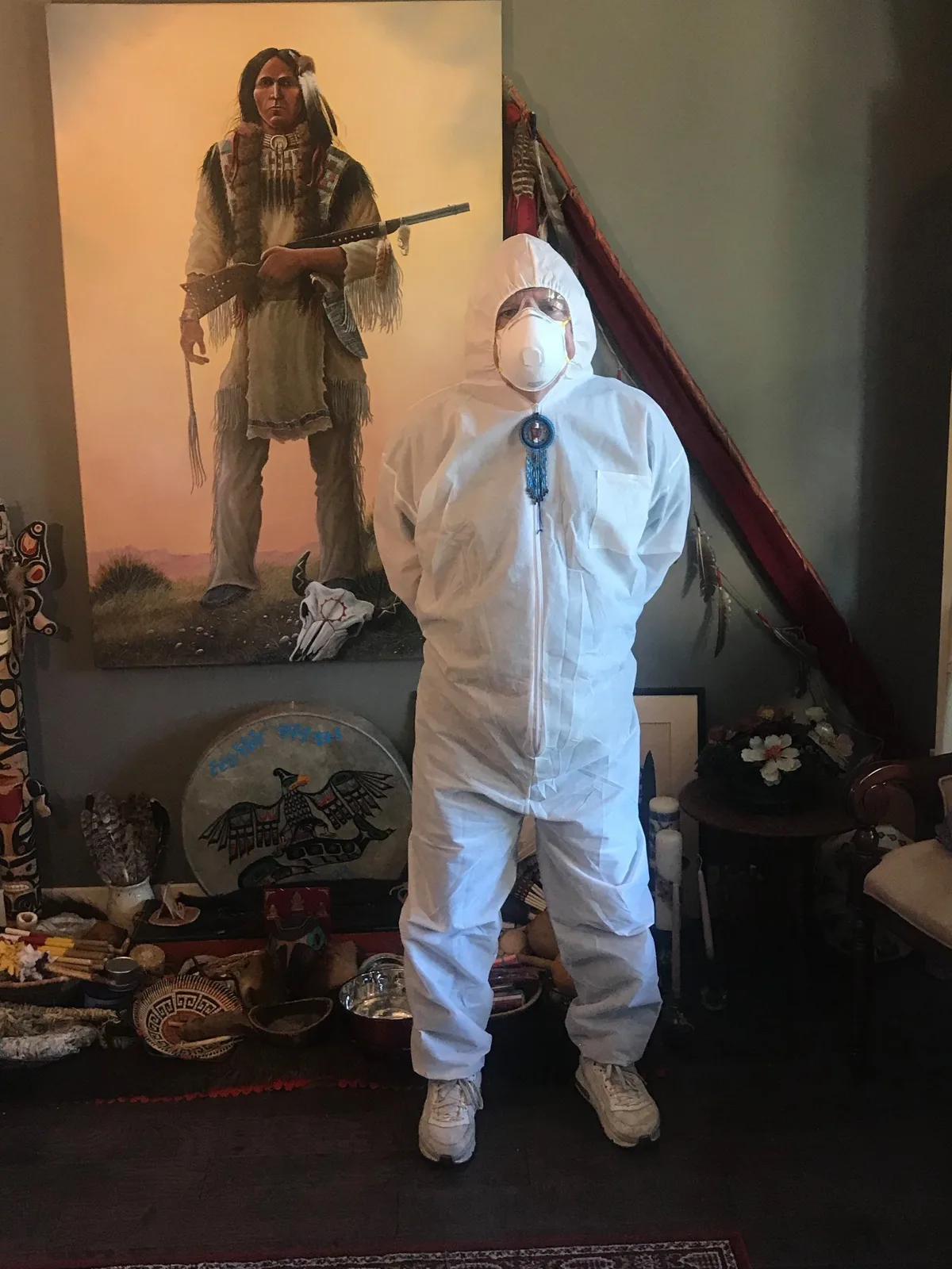 A person in a white hooded protective suit and N95 mask, wearing a blue beaded medallion, stands before a painting of a Plains warrior, a painted drum and ceremonial items.