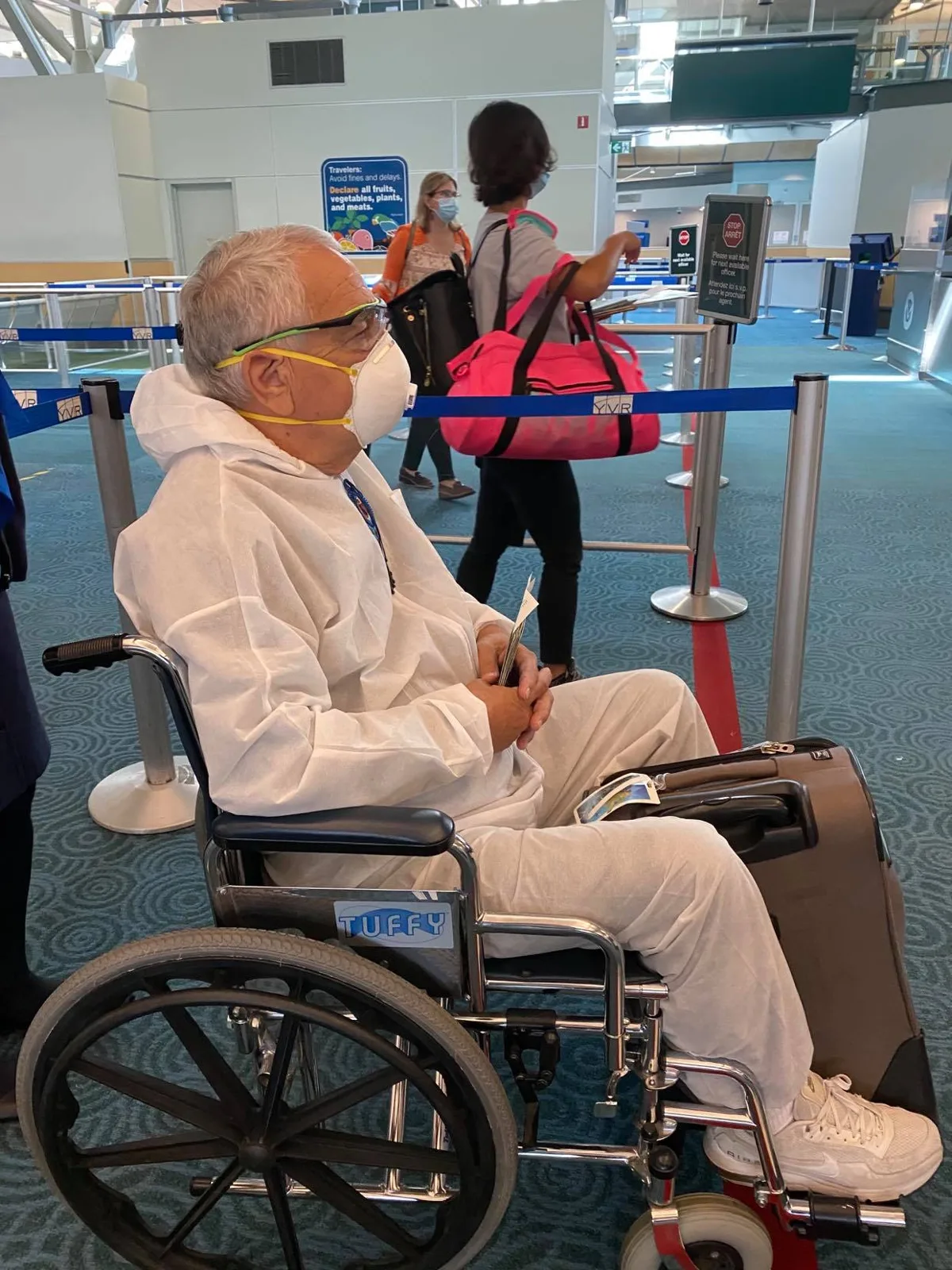 Phil Lane Jr., a grey-haired man in a white protective suit, face mask and glasses, sits in a wheelchair holding papers in an airport customs line, with masked travelers nearby.