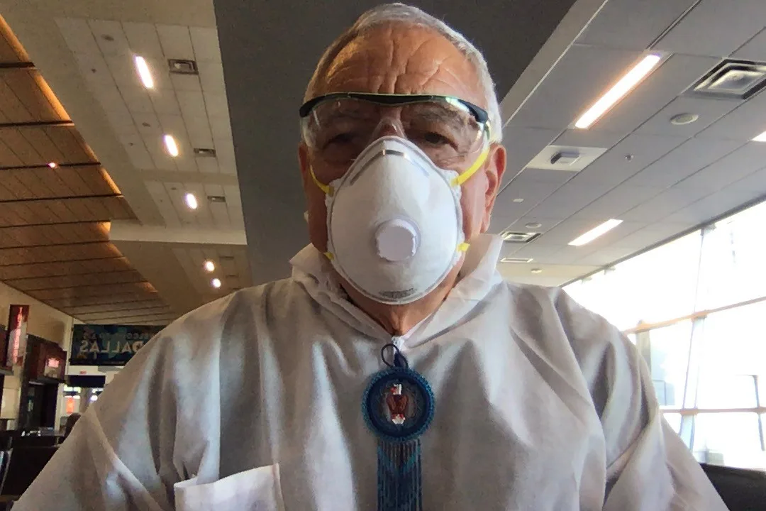 An elder in a white protective suit, N95 mask, safety goggles and a beaded blue medallion takes a selfie in a nearly empty airport terminal, a Dallas sign behind him.