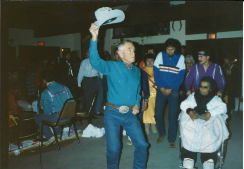 Phil Lane Jr.'s father, an older man in a blue shirt, jeans and silver belt buckle, dances holding a white cowboy hat aloft, with people and an elder in a wheelchair behind him.