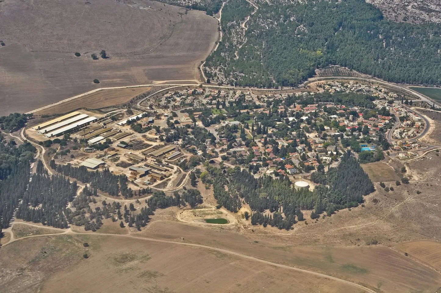 Aerial view of a rural settlement surrounded by dry brown fields and pine forest, with clustered red-roofed houses and long farm buildings.