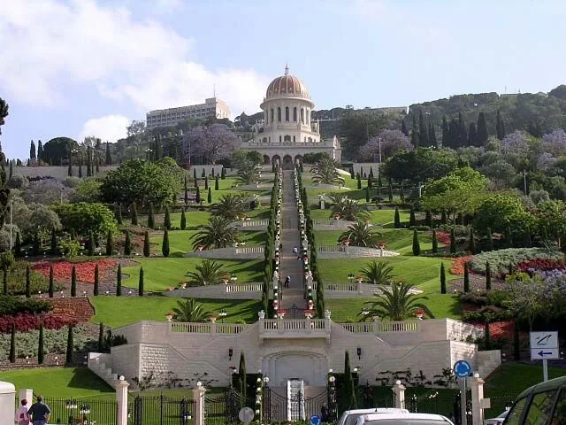 Terraced gardens climbing Mount Carmel toward a golden-domed shrine, with a central stairway lined by cypresses, palms and flower beds.