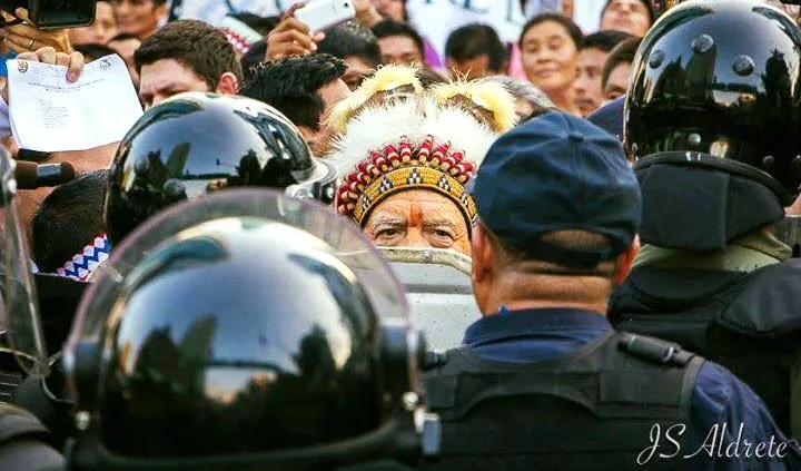 An elder in a white feathered headdress with a beaded band stands face to face with helmeted riot police in a crowd, photographers and onlookers behind him.
