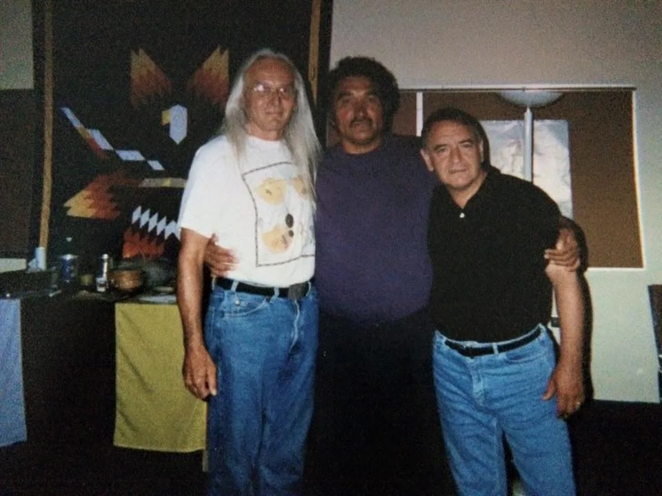 Three men stand arm in arm, Phil Lane Jr. with Sequoia Trueblood and Dan Highway, before a star quilt wall hanging at a 1992 workshop in Winnipeg.