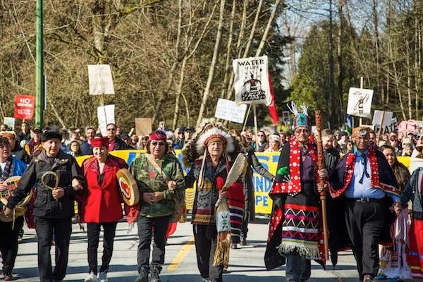 Indigenous leaders in regalia, one in a feather headdress and others in button blankets, lead a large march down a road with hand drums and signs including "Water Is Life."
