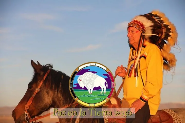 An elder in a feathered war bonnet and yellow shirt rides a brown horse at sunset. A circular emblem shows a white buffalo, with the words "Paz en la Tierra 2030."