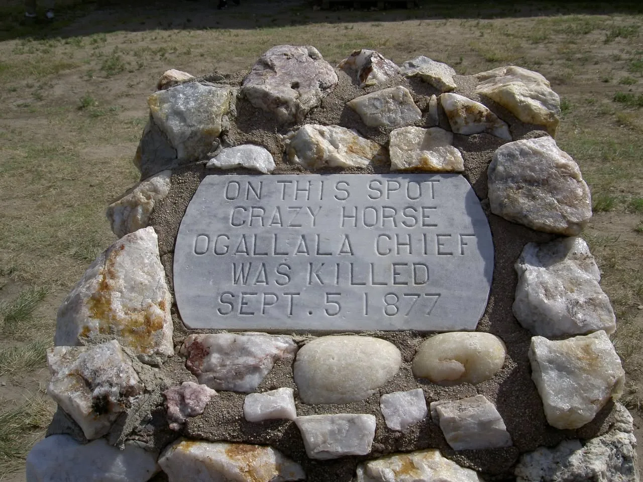 A white marble plaque set in a cairn of quartz stones reads: "On this spot Crazy Horse Ogallala Chief was killed Sept. 5 1877," on dry grassy ground.