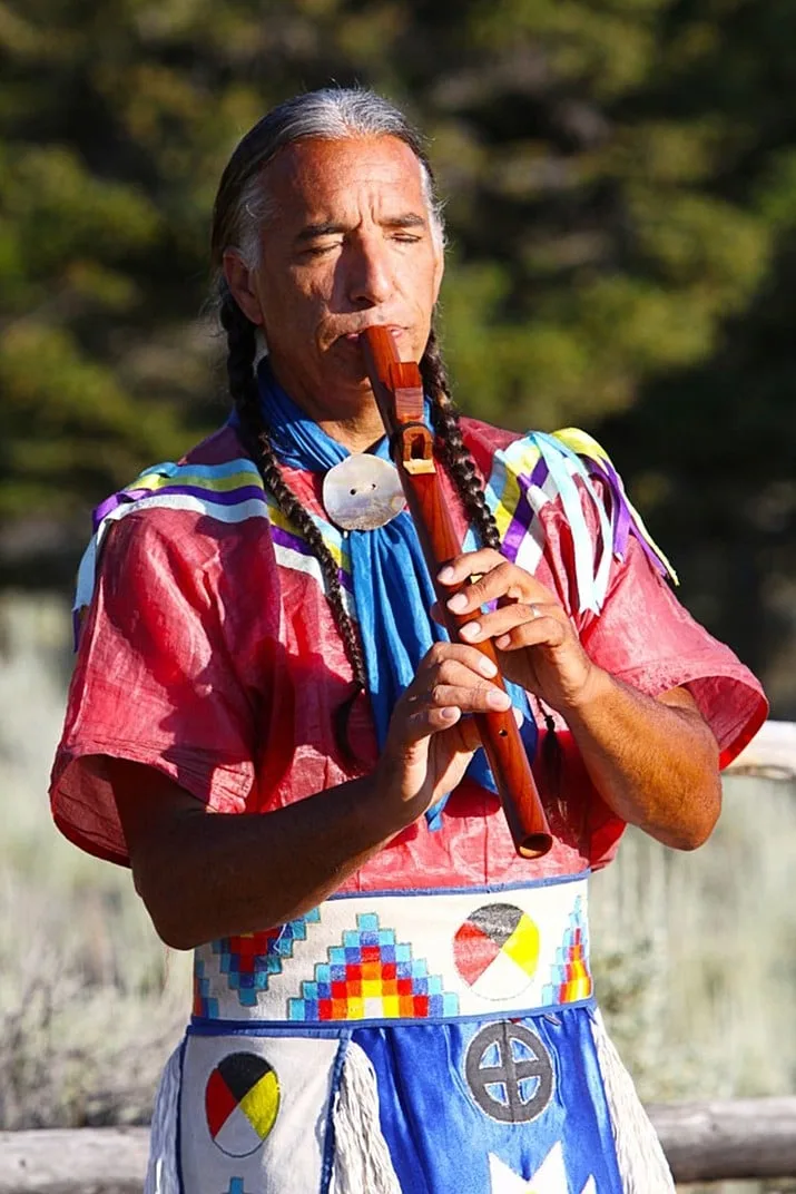 A man with long gray braids, wearing a red shirt with ribbons and a beaded belt with medicine wheel designs, plays a wooden flute with eyes closed.