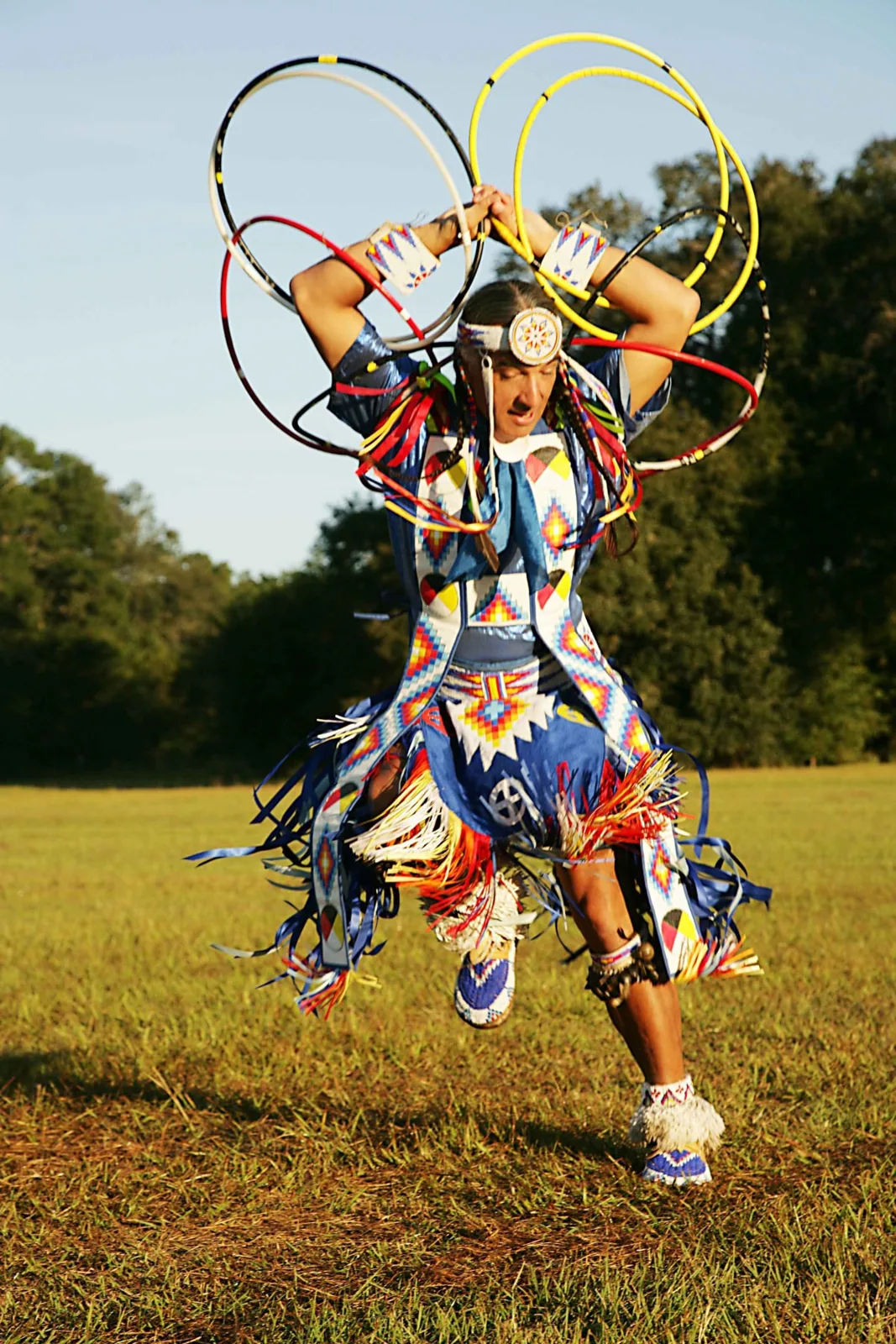A hoop dancer in bright blue beaded regalia with colorful fringe holds many red, yellow, white and black hoops overhead while dancing on a field.