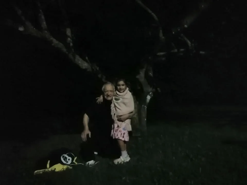 A white-haired elder kneels on the grass at the base of a large tree at night, his arm around a young girl in a shawl standing beside him.