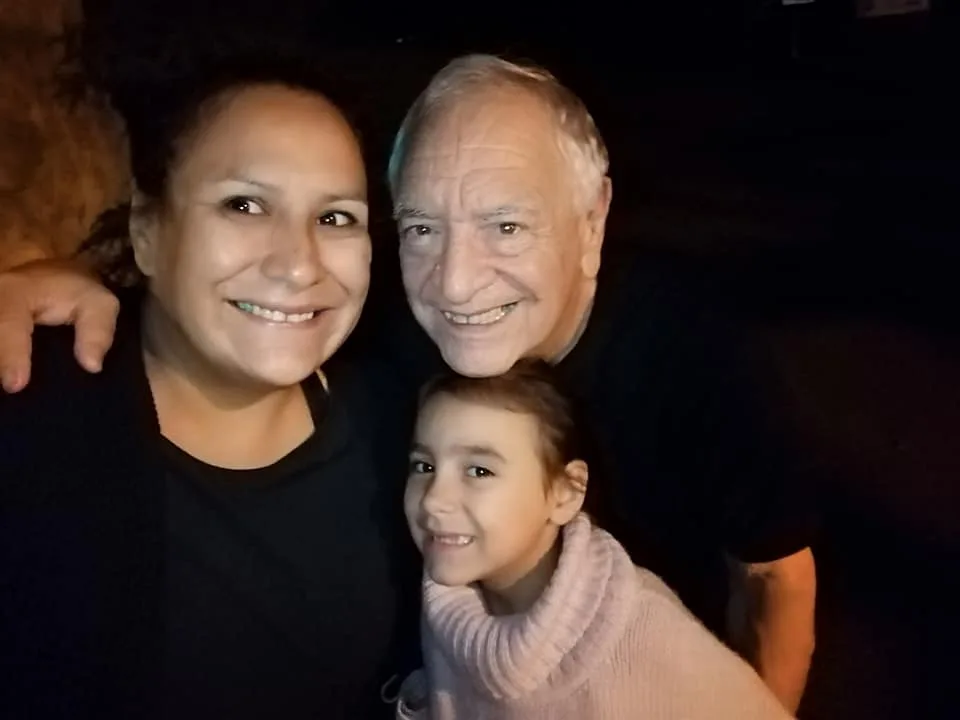 A smiling woman and a white-haired elder lean together at night, with a young girl in a pink sweater tucked between them.